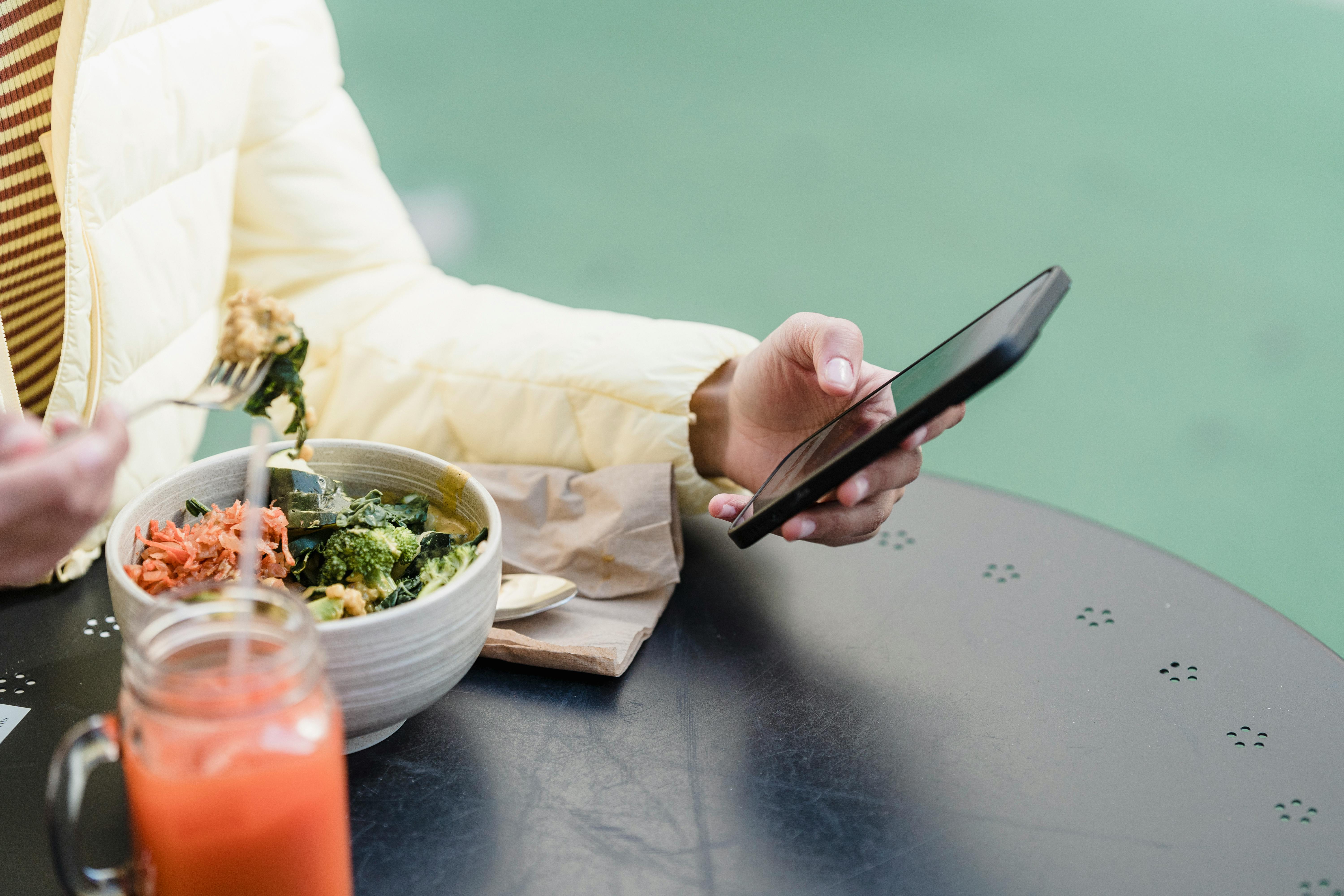 Crop unrecognizable female with cellphone sitting at cafe table with delicious vegan salad and smoothie in daylight
